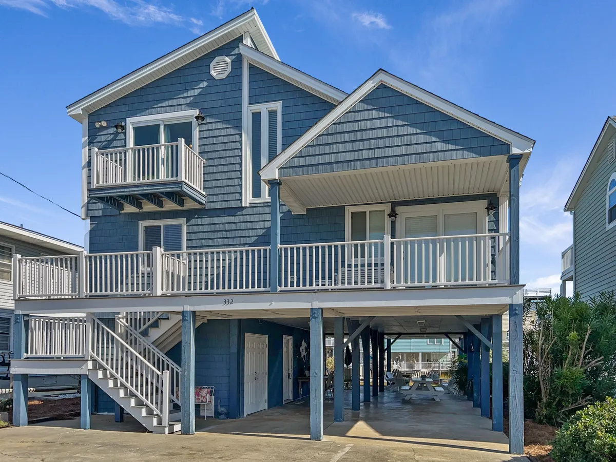The exterior of a pet-friendly beach house, featuring blue siding, balconies, a staircase, and an underneath parking area. The space is suitable for family gatherings and outdoor activities, with open areas ideal for pets to roam safely and for guests to enjoy the porch or yard.