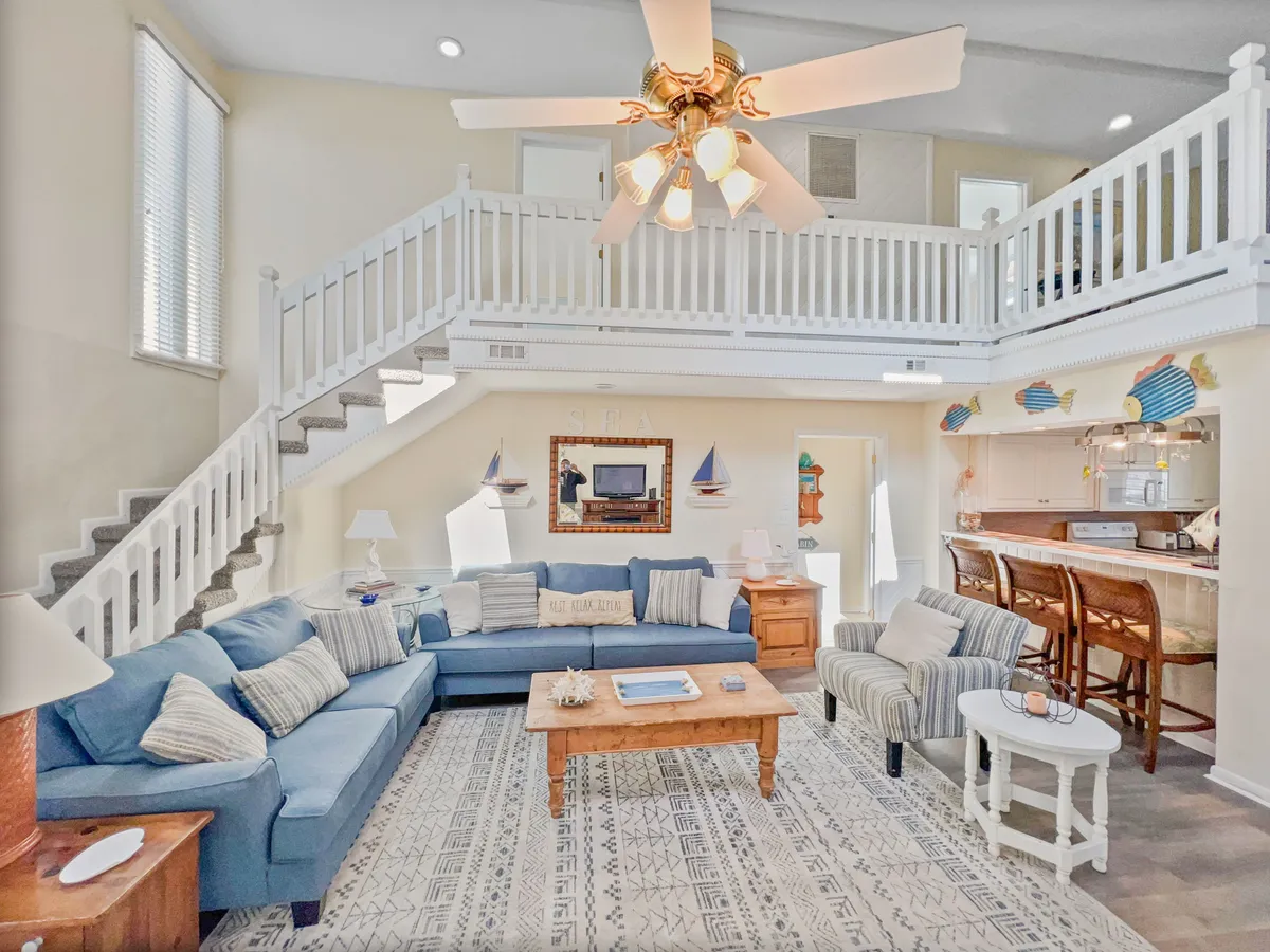 A spacious and casual living area with a large blue sectional sofa and a wooden coffee table sits adjacent to a staircase leading to a loft. The area is organized for relaxation, socializing, and entertaining, with plenty of space for group activities such as games and movie marathons.