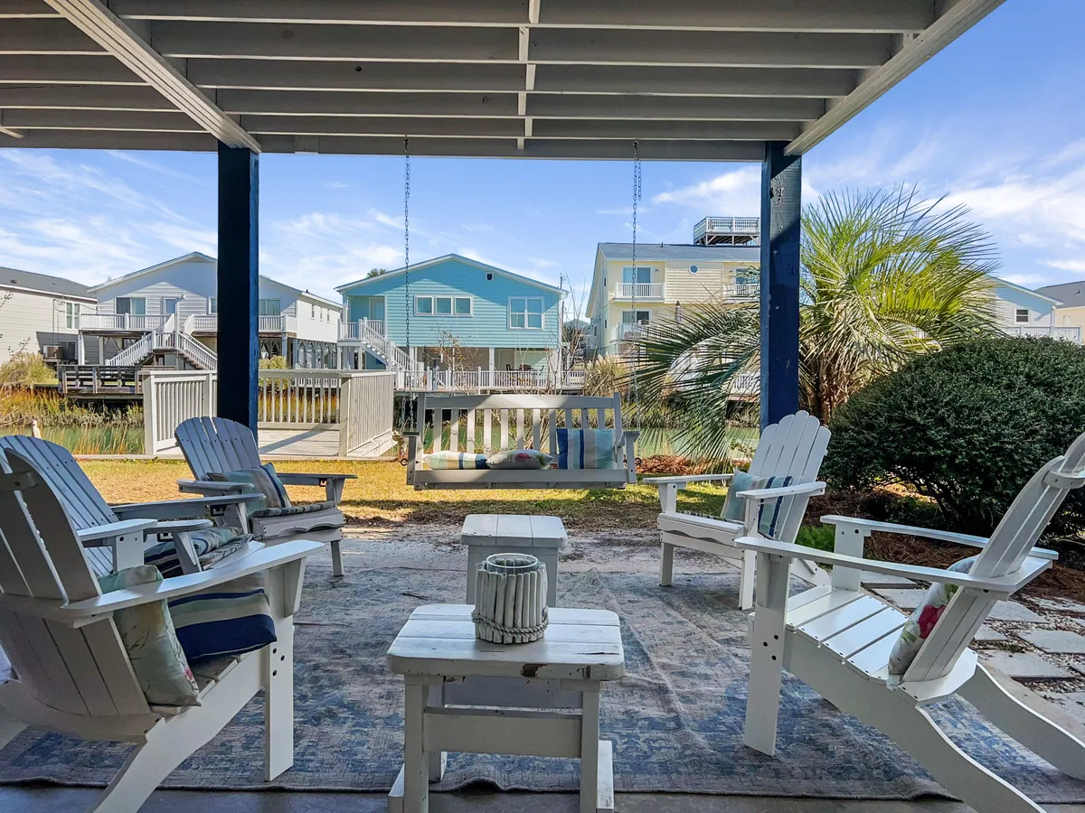 The image shows the tranquil exterior of a spacious beach house, featuring white wooden chairs and a charming swing bench on the porch, framed by swaying palm trees. The porch overlooks serene channel views, with neighboring blue houses providing a classic coastal backdrop. The inviting setting suggests opportunities for relaxing with morning coffee, gathering with loved ones, or simply soaking in North Myrtle Beach’s breezy ambiance.
