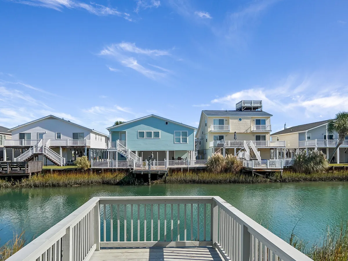 A tranquil exterior view of waterfront houses lining a serene canal, with a dock extending over the water and calm reflections on the surface. The setting evokes the peaceful, laid-back lifestyle associated with North Myrtle Beach, highlighting access to water-based activities and picturesque walks.
