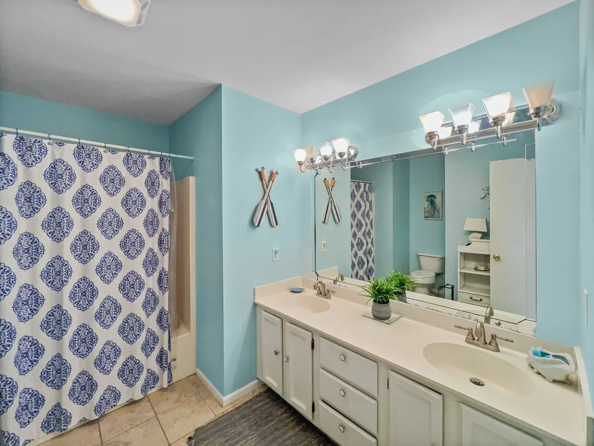 A bathroom interior with a double sink vanity, blue walls, a decorative shower curtain, and mirrored lighting fixtures. The space feels both functional and soothing, ideal for multiple guests getting ready at once.