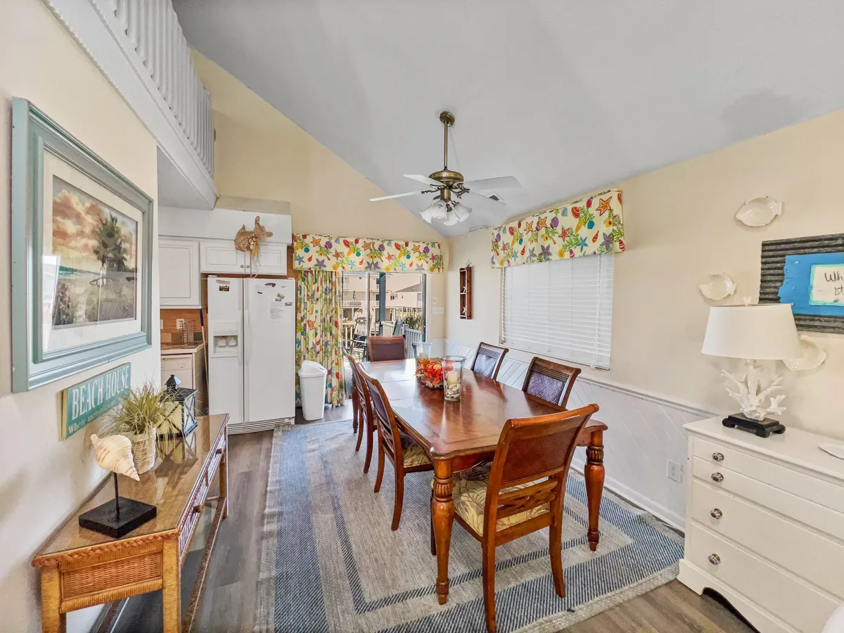 A welcoming dining area featuring a large table, colorful window treatments, and beach-themed artwork. The setup invites guests to gather for shared meals, casual chats, or group celebrations, further enhancing the sense of togetherness.