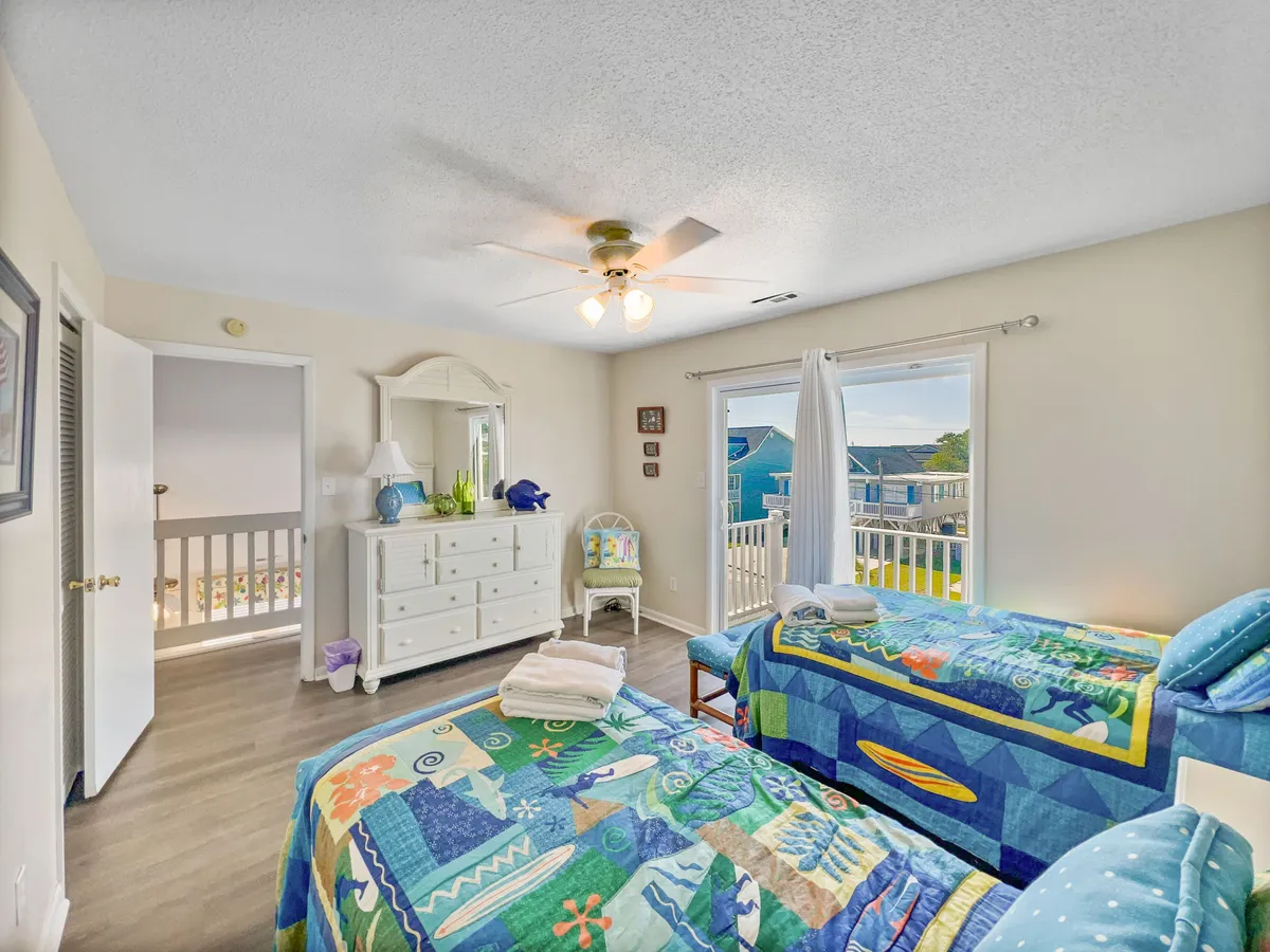This room includes two beds with colorful bedding, a dresser with a mirror, large windows flooding the space with light, and a small chair and table. The space feels playful yet restful, making it ideal for children or friends sharing a room.